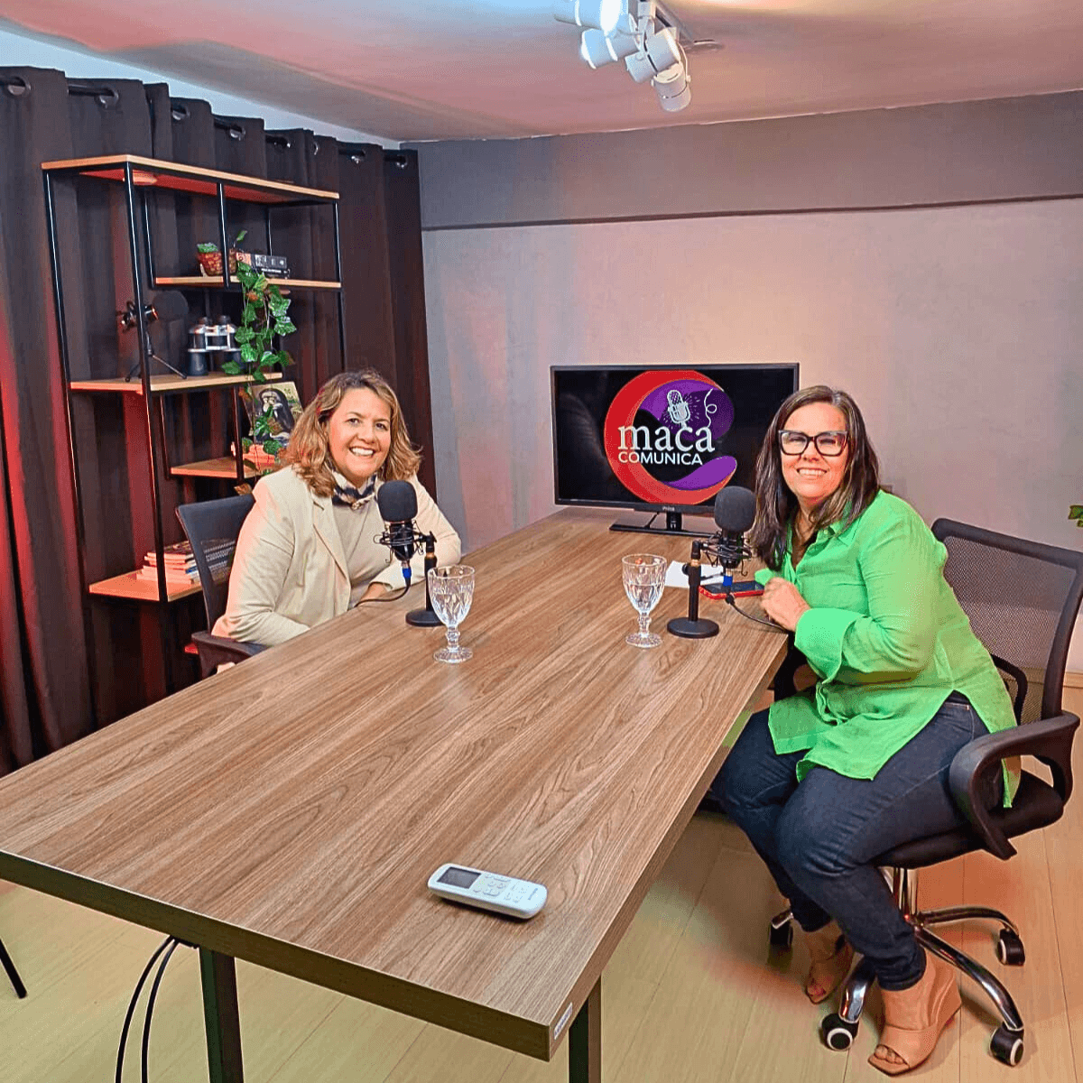 Duas mulheres sentadas em uma mesa com microfones em um estúdio de podcast.