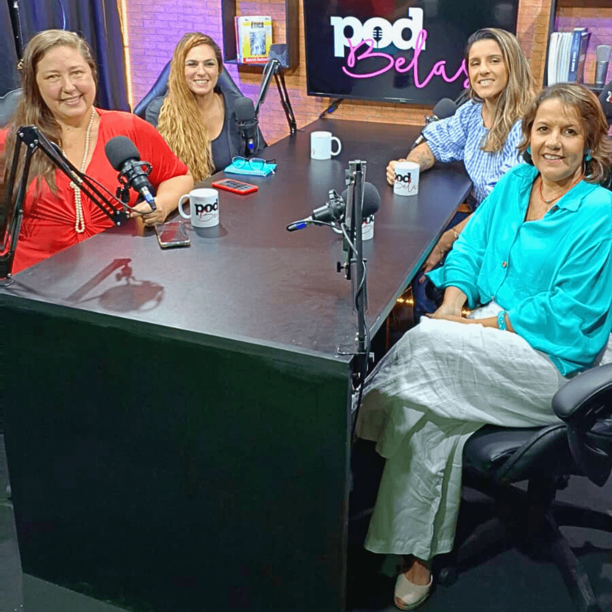 Três mulheres sentadas ao redor de uma mesa com microfones, gravando um podcast.