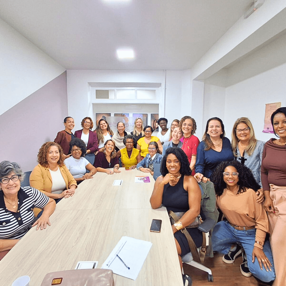 Grande grupo de mulheres reunidas, algumas em pé e outras sentadas, em uma sala iluminada.